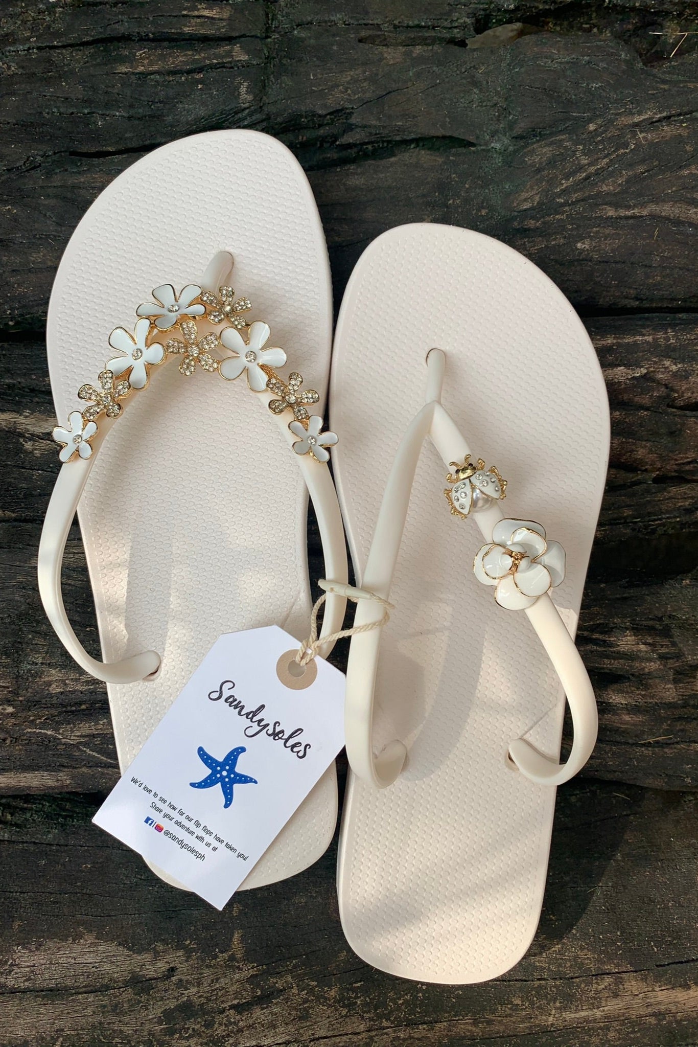 Pair of white flipflops with floral embellishments, featuring a SandySoles brand tag.