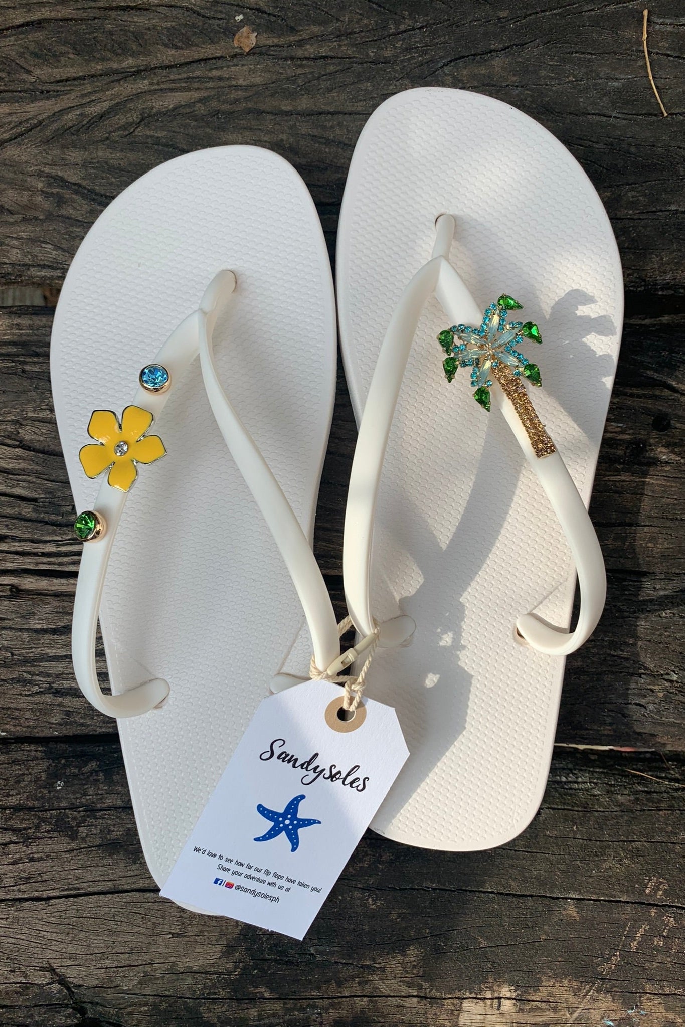 White flip-flops with decorative charms on straps, featuring a 'SandySoles' tag.