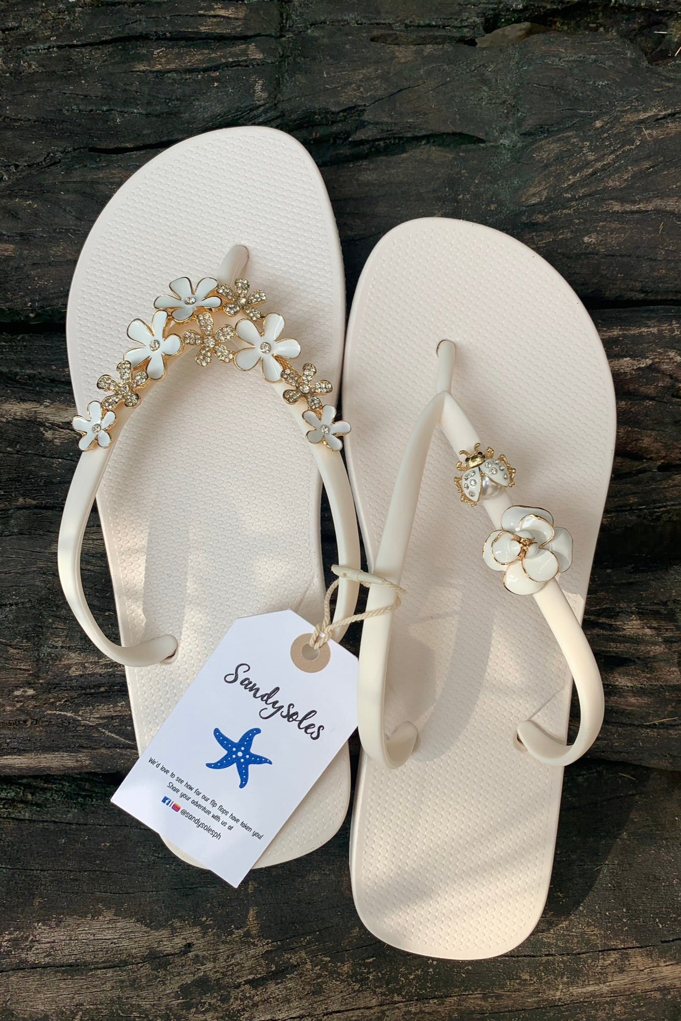 Pair of white flipflops with a beetle and floral decorative charms, featuring a SandySoles brand tag.