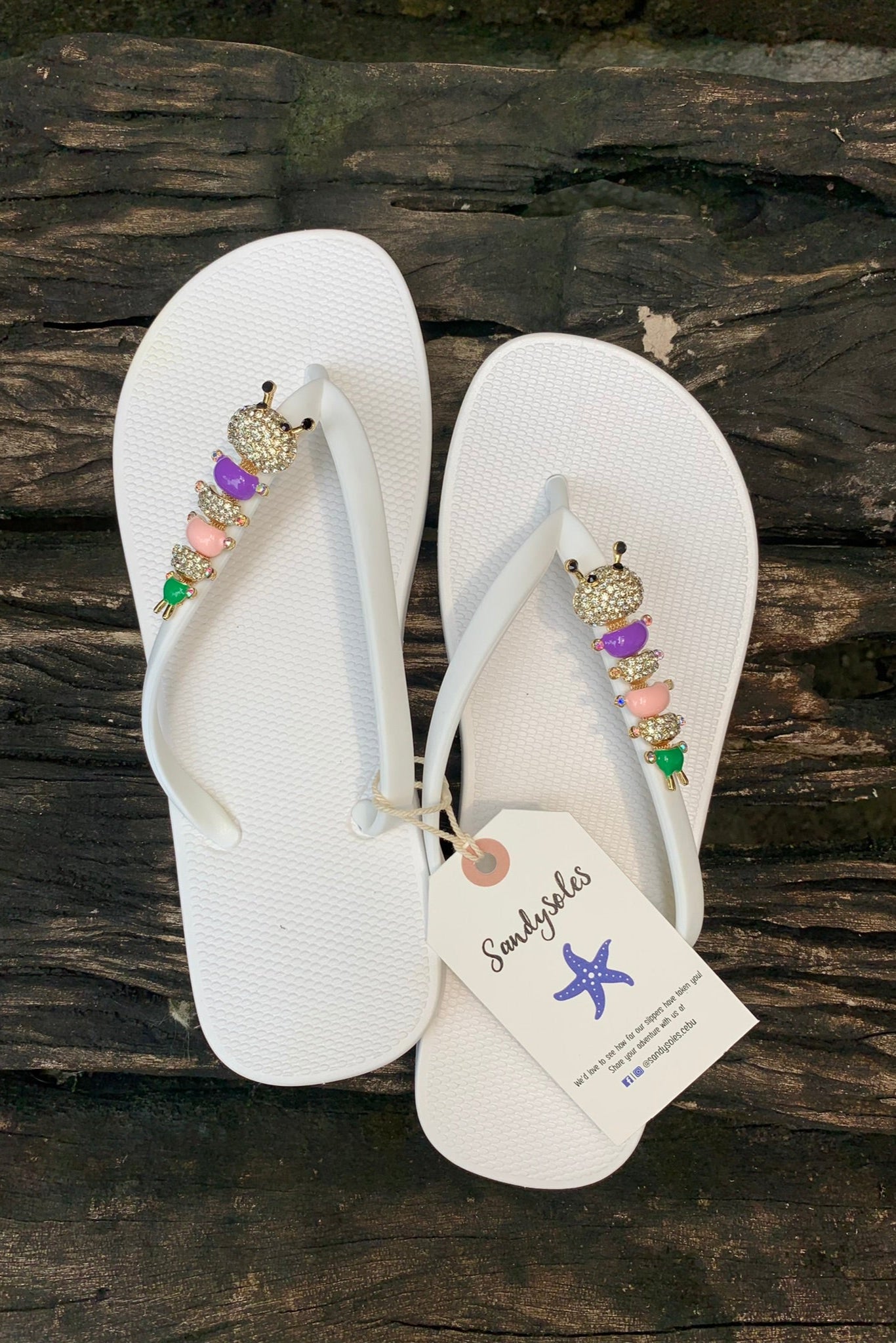 White flip-flops with decorative caterpillar charms, featuring a sandysoles tag