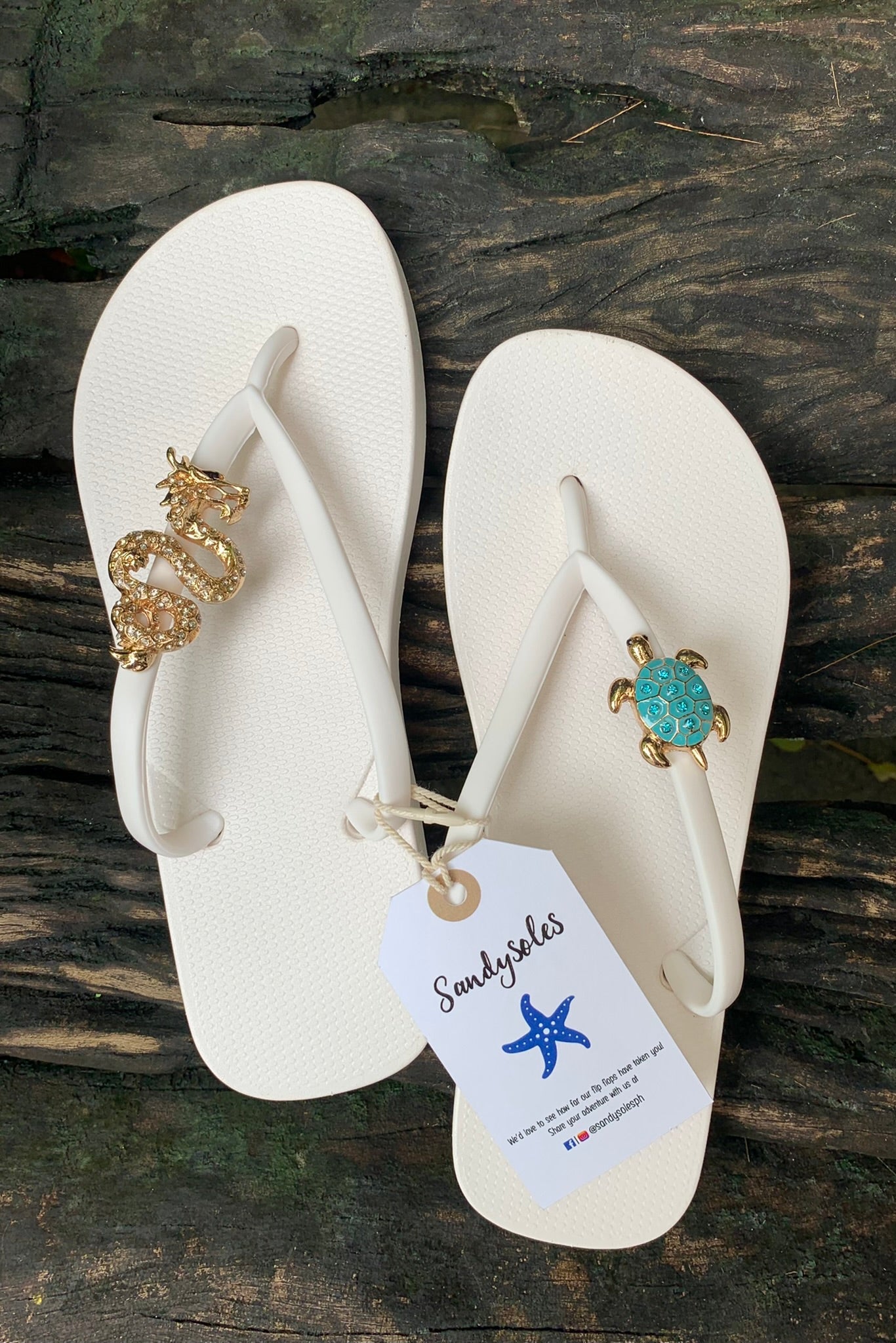 White flip-flops with decorative dragon and turtle charms, featuring a Sandysoles tag