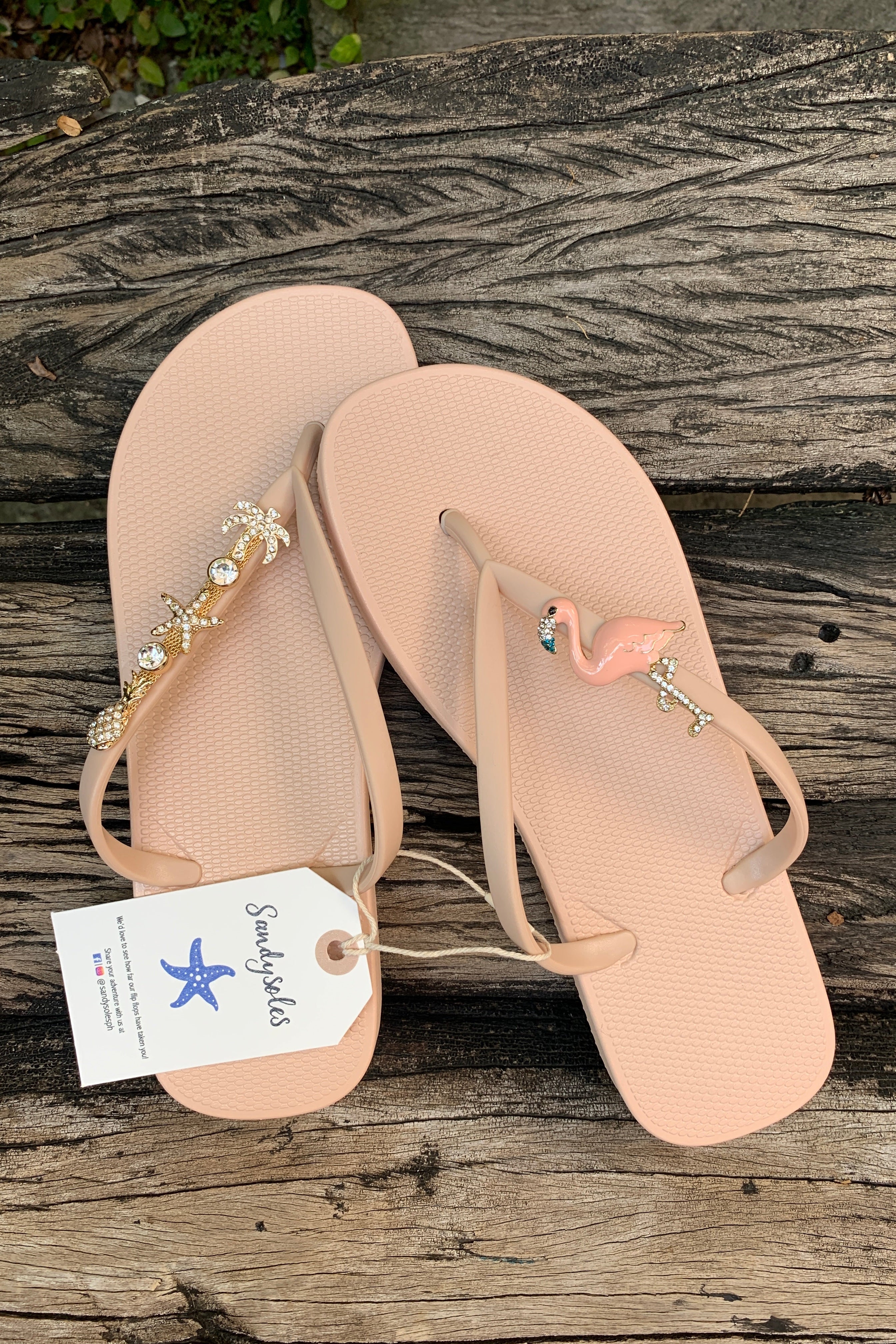 Pair of beige flip-flops with decorative flamingo and gemstone charms, featuring a 'SandySoles' tag.