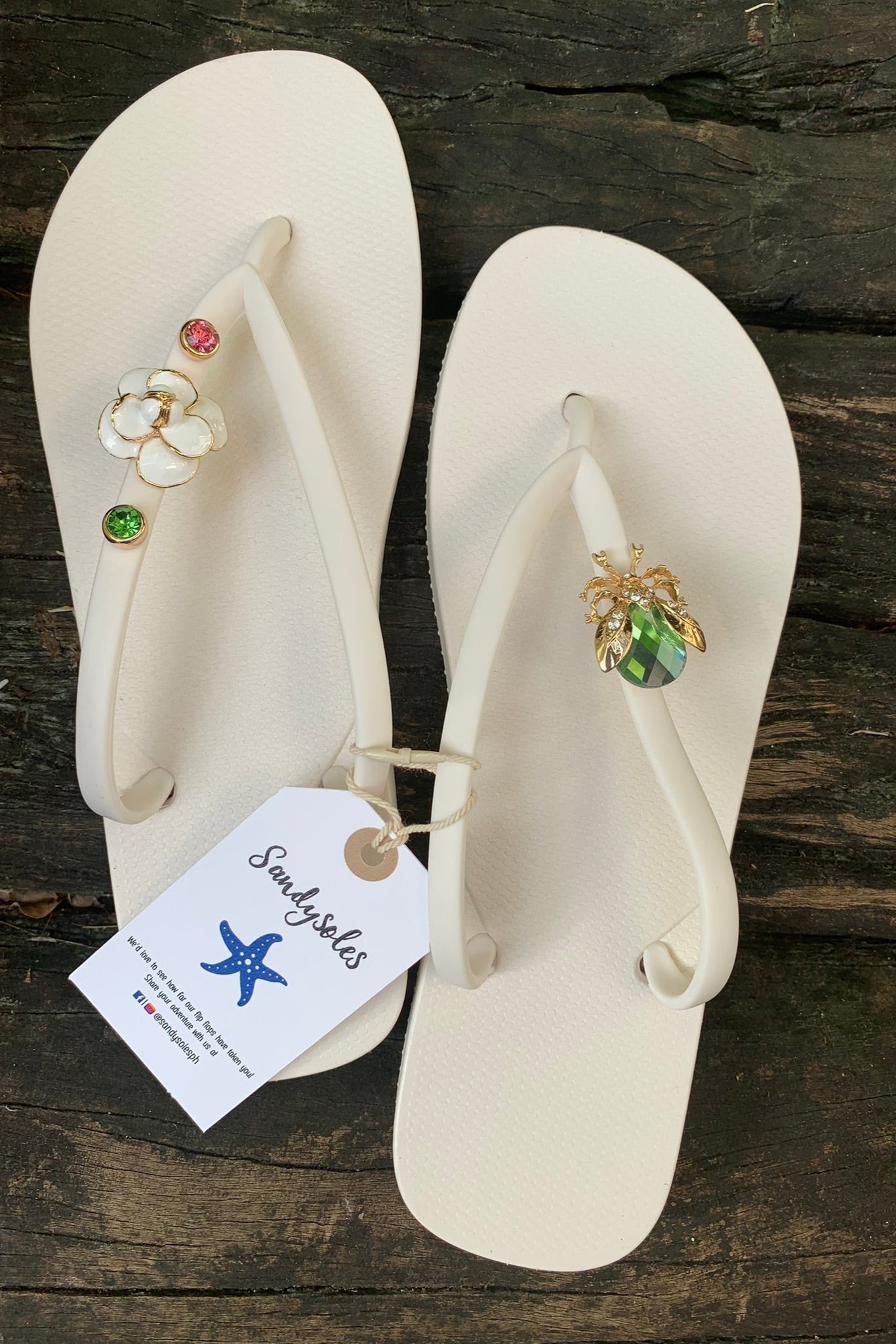 Pair of white flip-flops with gemstone and beetle charms on straps, featuring a 'Sandysoles' tag.