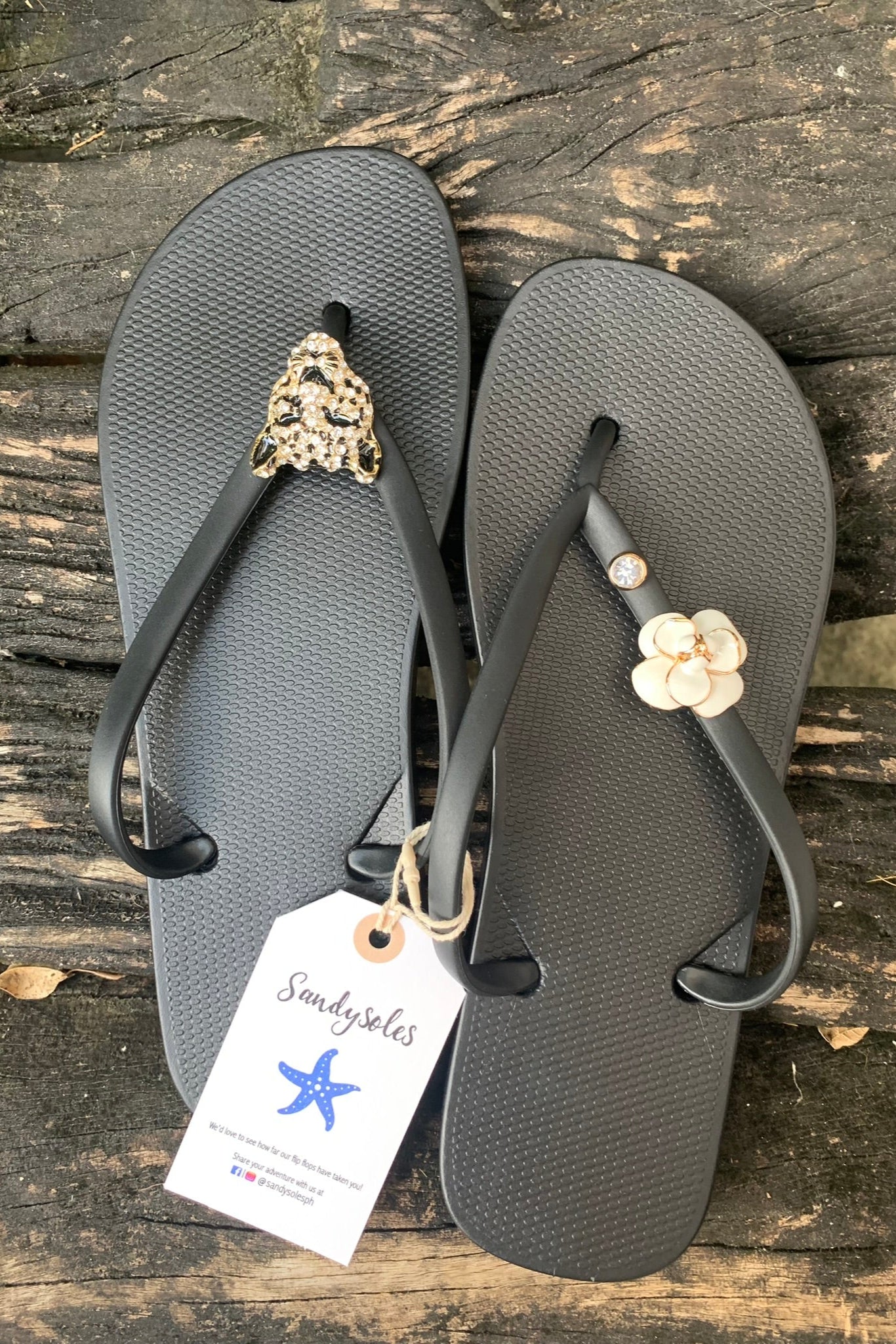black flip-flops with decorative jaguar, gemstone and camellia charms, featuring a 'Sandysoles' tag.