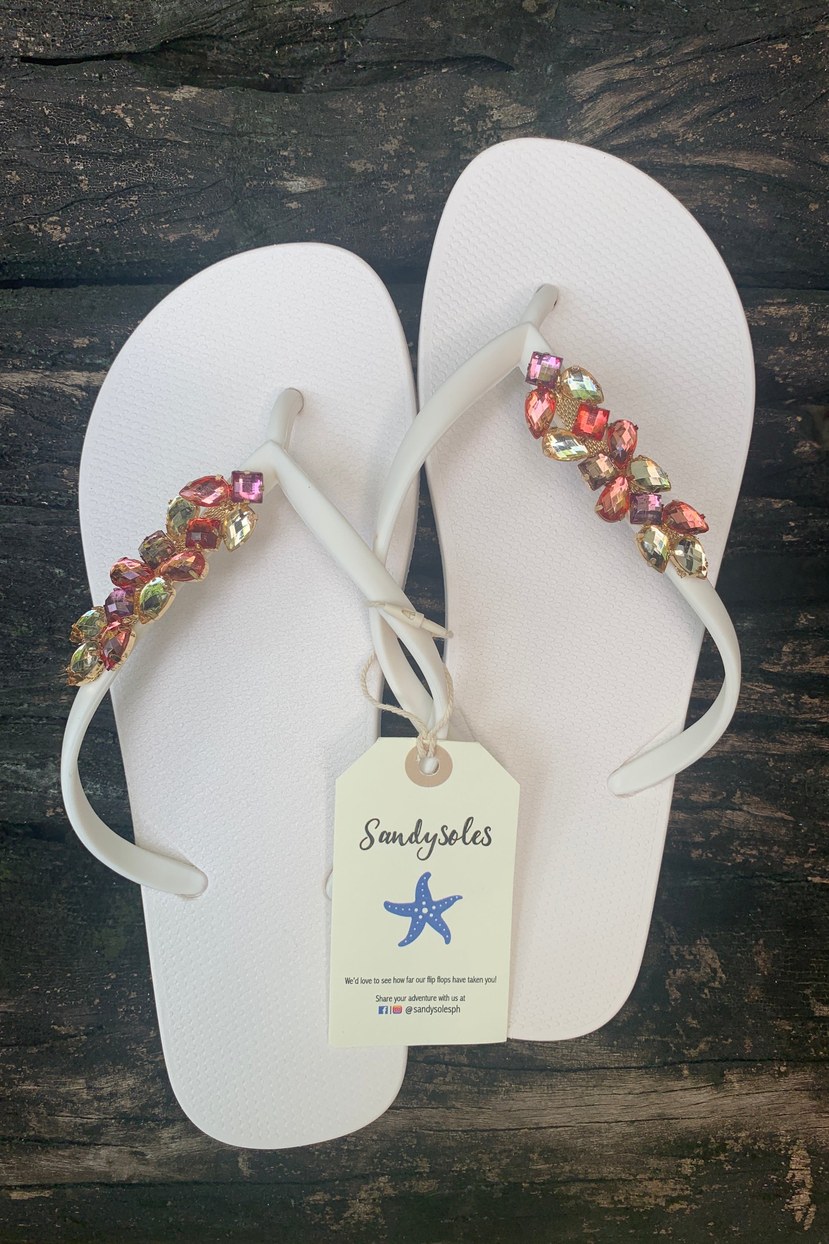 a pair of white flip-flops with multicolored gemstone leaf-shaped charm