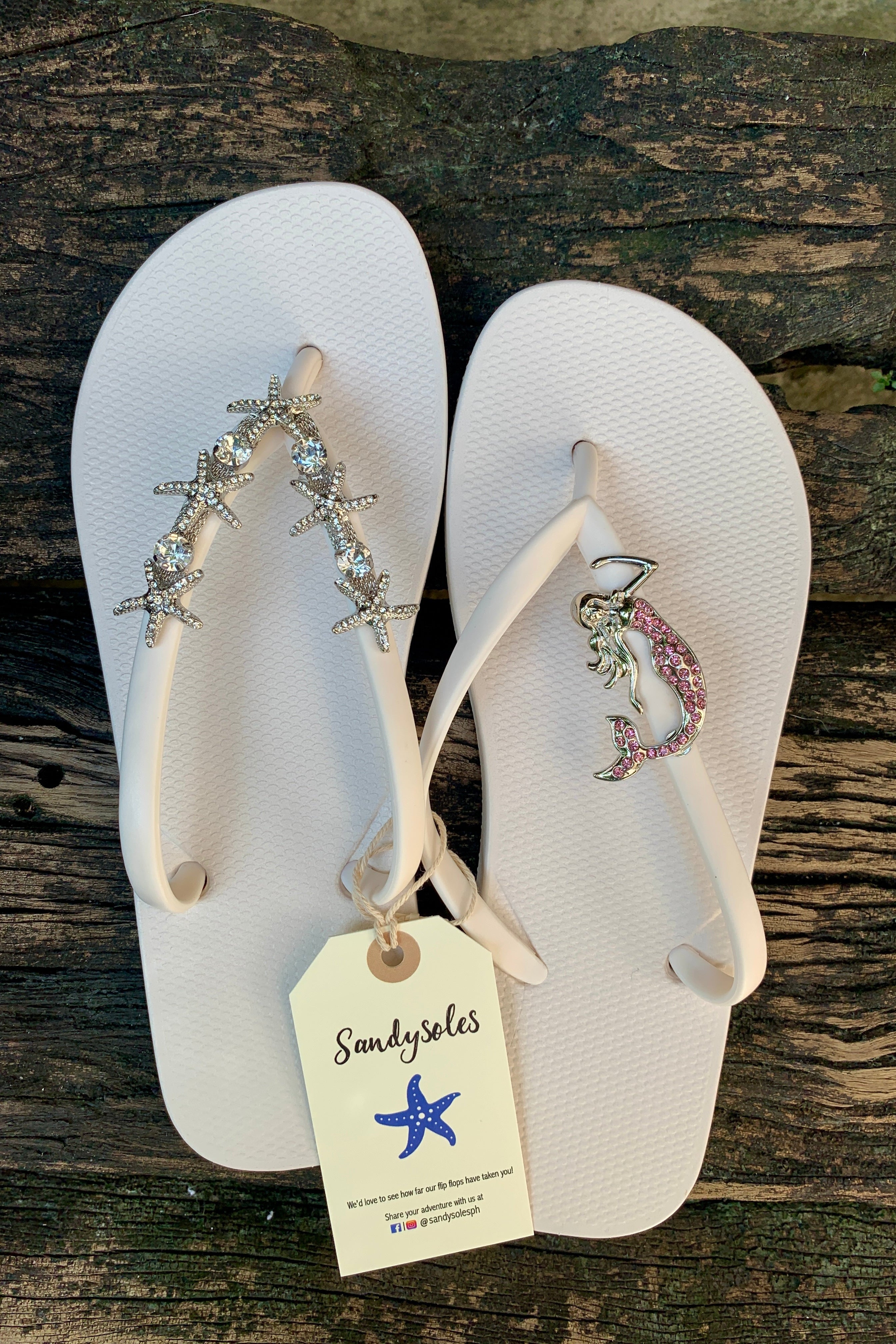 White flip-flops with mermaid and starfish decorative charms on straps, featuring a 'SandySoles' tag.