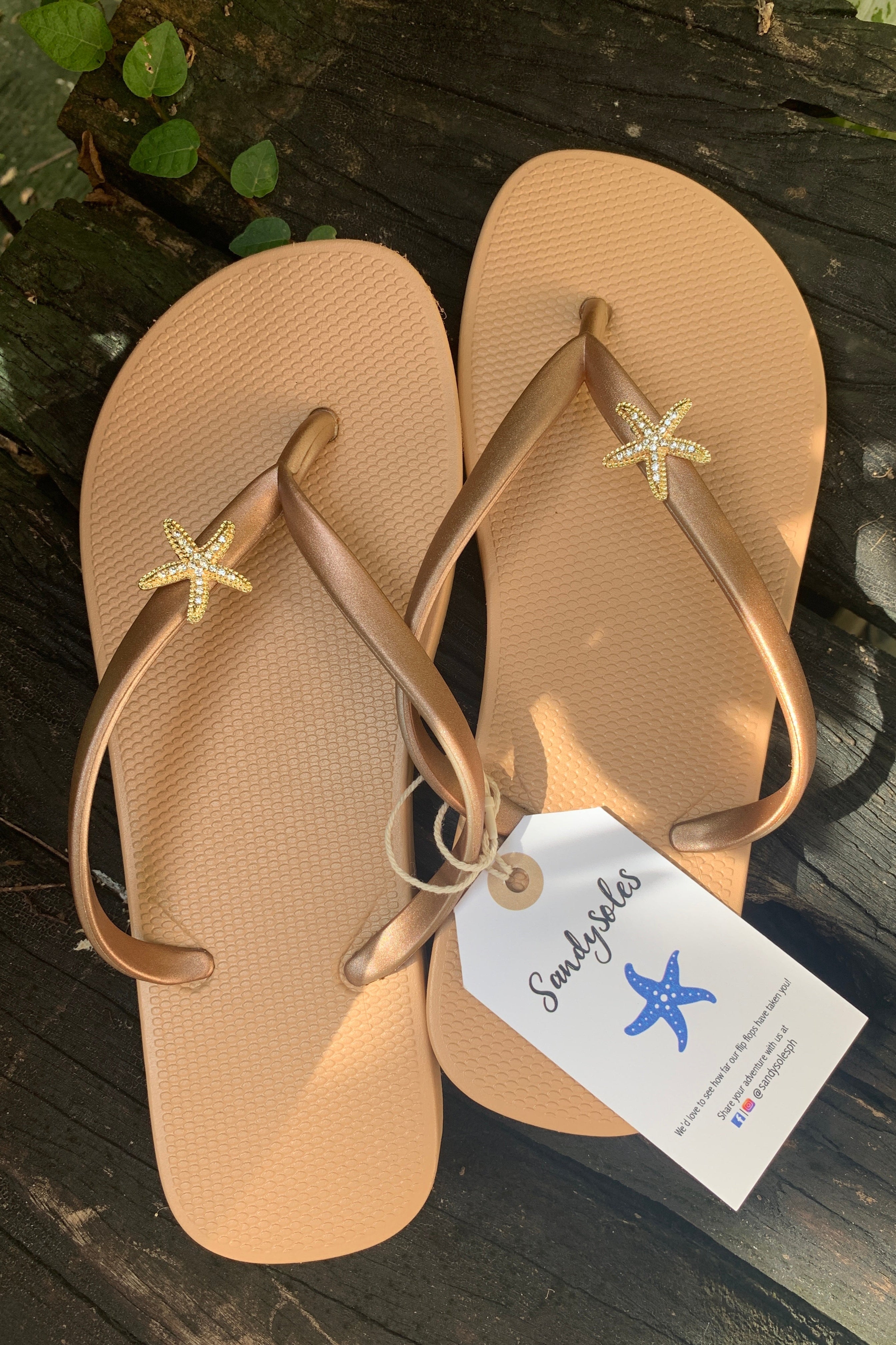 Beige flip-flops with starfish design charms on straps, featuring a 'Sandysoles' tag.