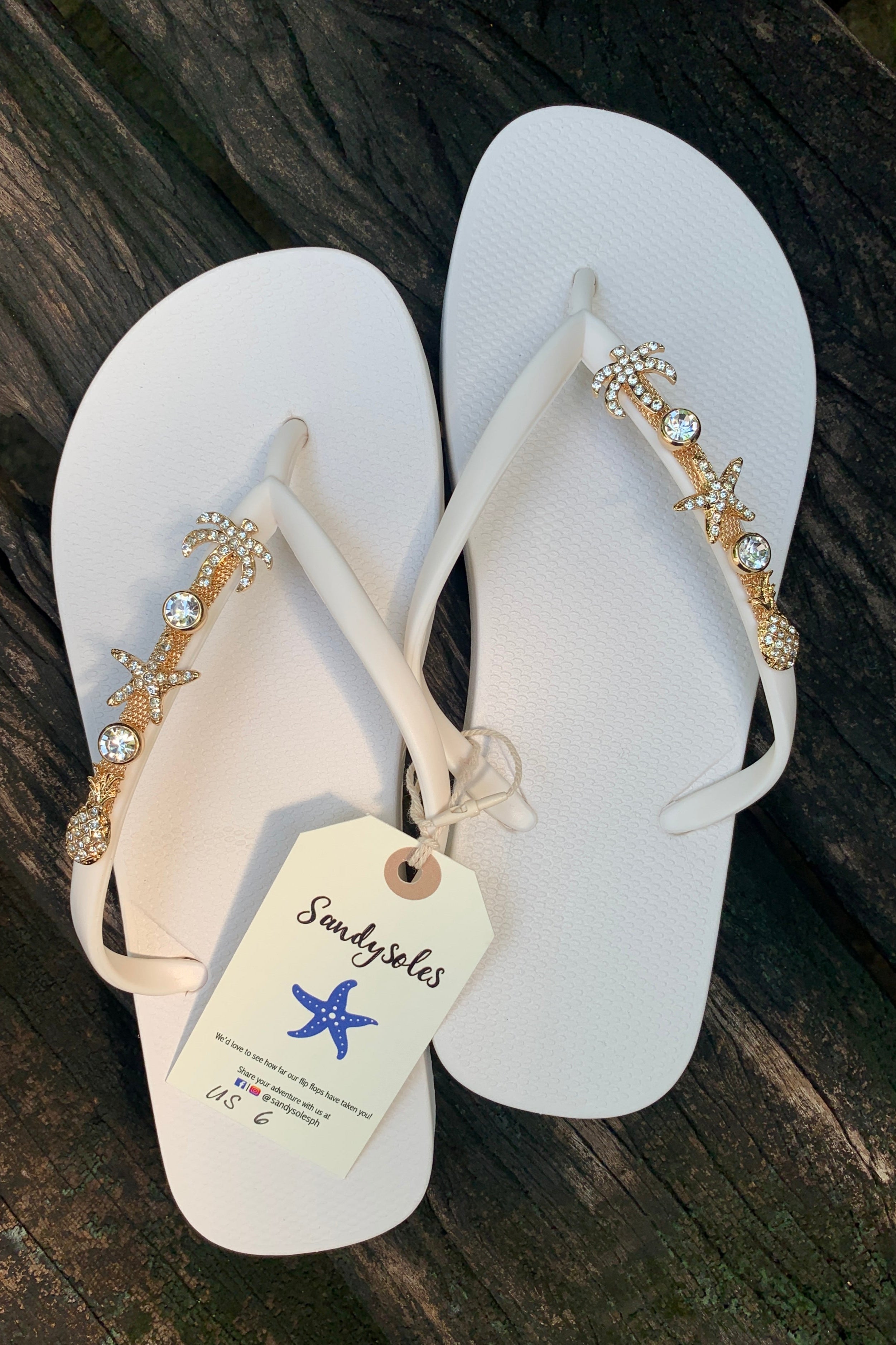 White flip-flops with decorative charm with palm tree, starfish, and pineapple design, featuring a 'Sandysoles' tag.