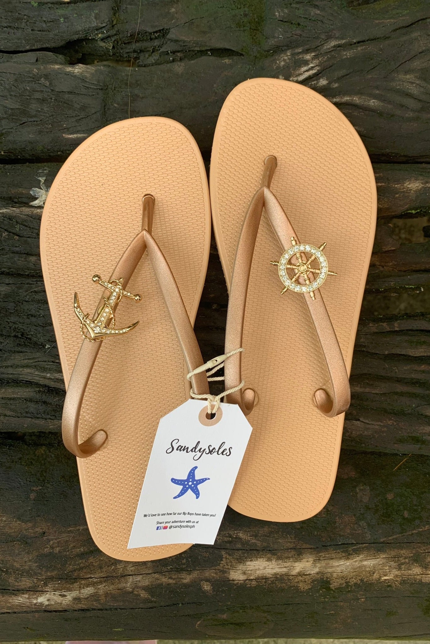 Pair of nude/tan flip-flops with anchor and compass design on a wooden surface, featuring a Sandysoles tag.