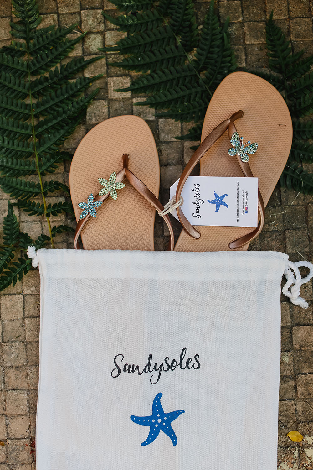 Pair of tan flip-flops with starfish decorations on a 'Sandysoles' branded drawstring bag.