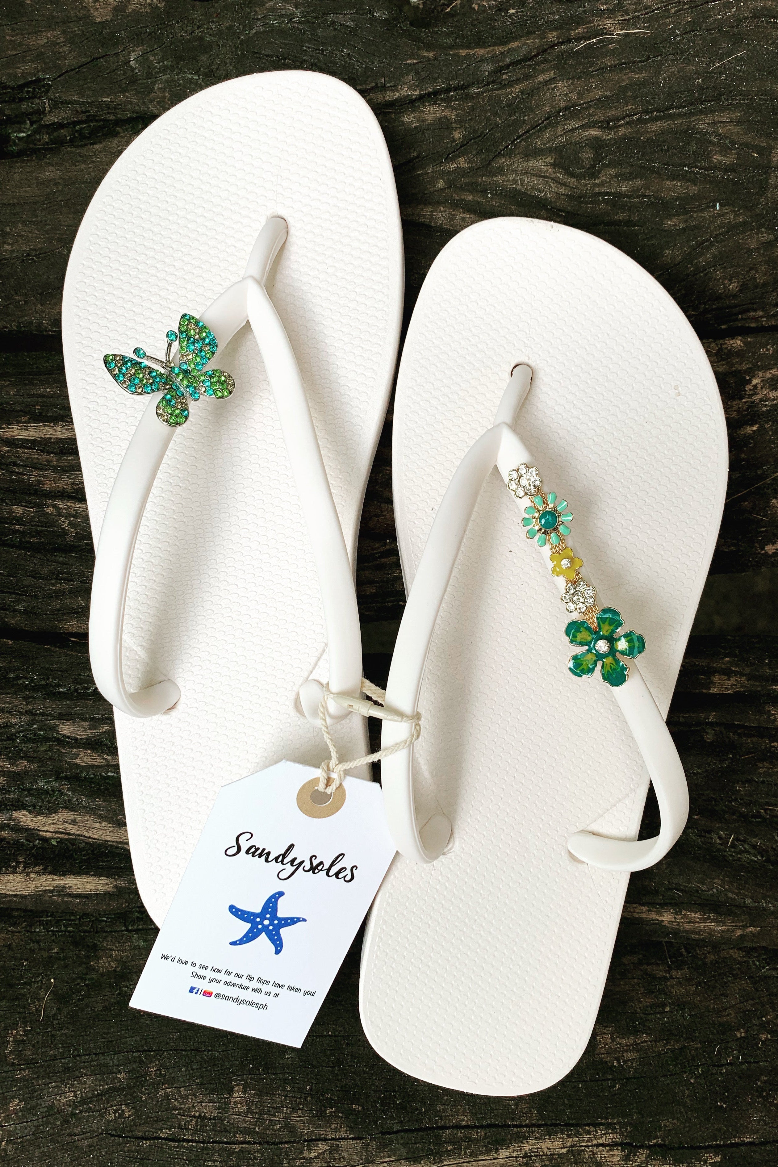 White flip-flops with decorative charms on straps, featuring the brand 'Sandysoles'.