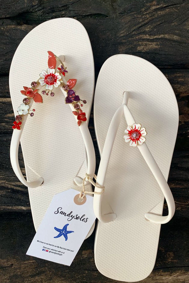 White flip-flops with decorative floral charms, featuring a SandySoles tag