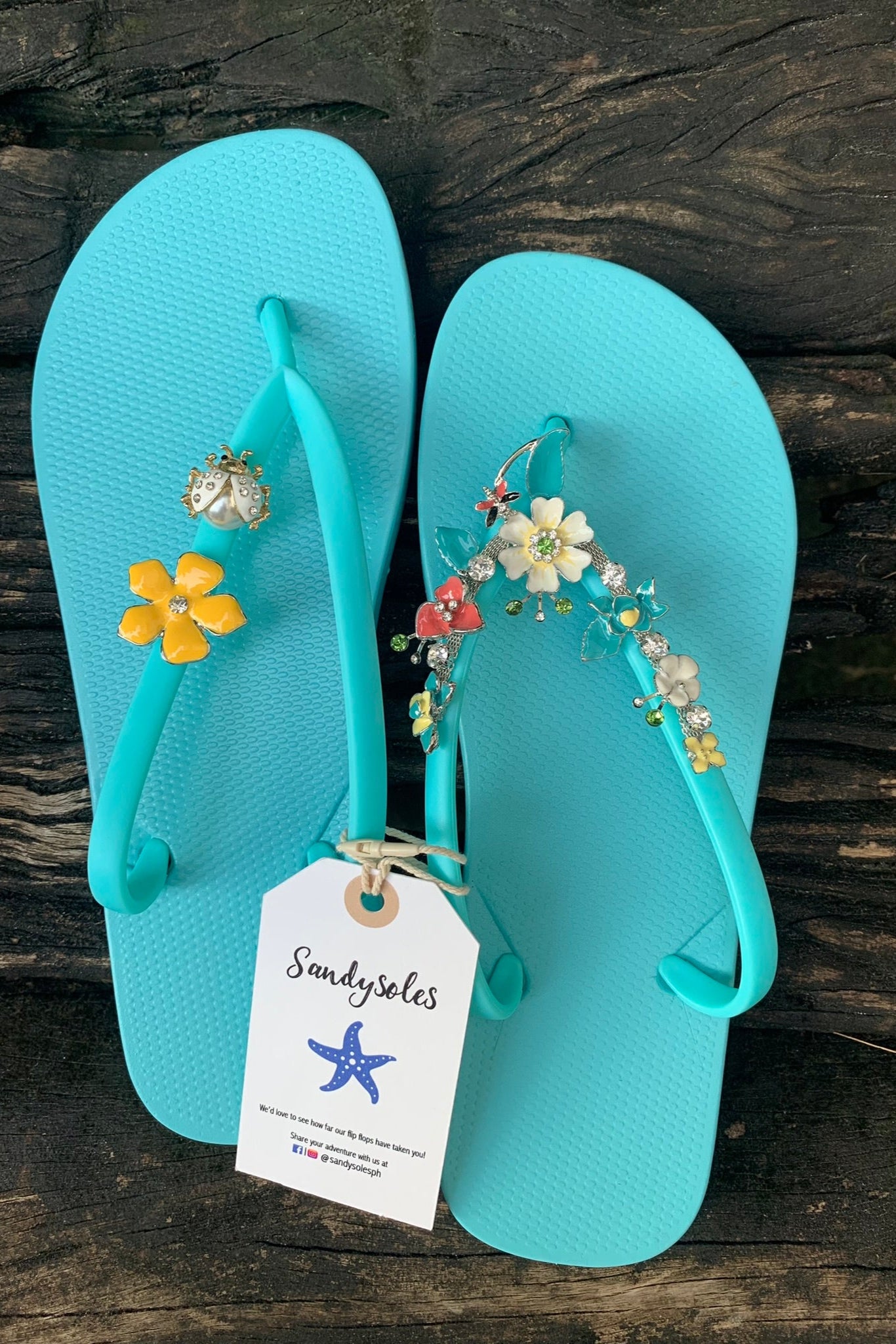 Pair of aqua flip-flops with floral and beetle charms, featuring a 'Sandysoles' tag.