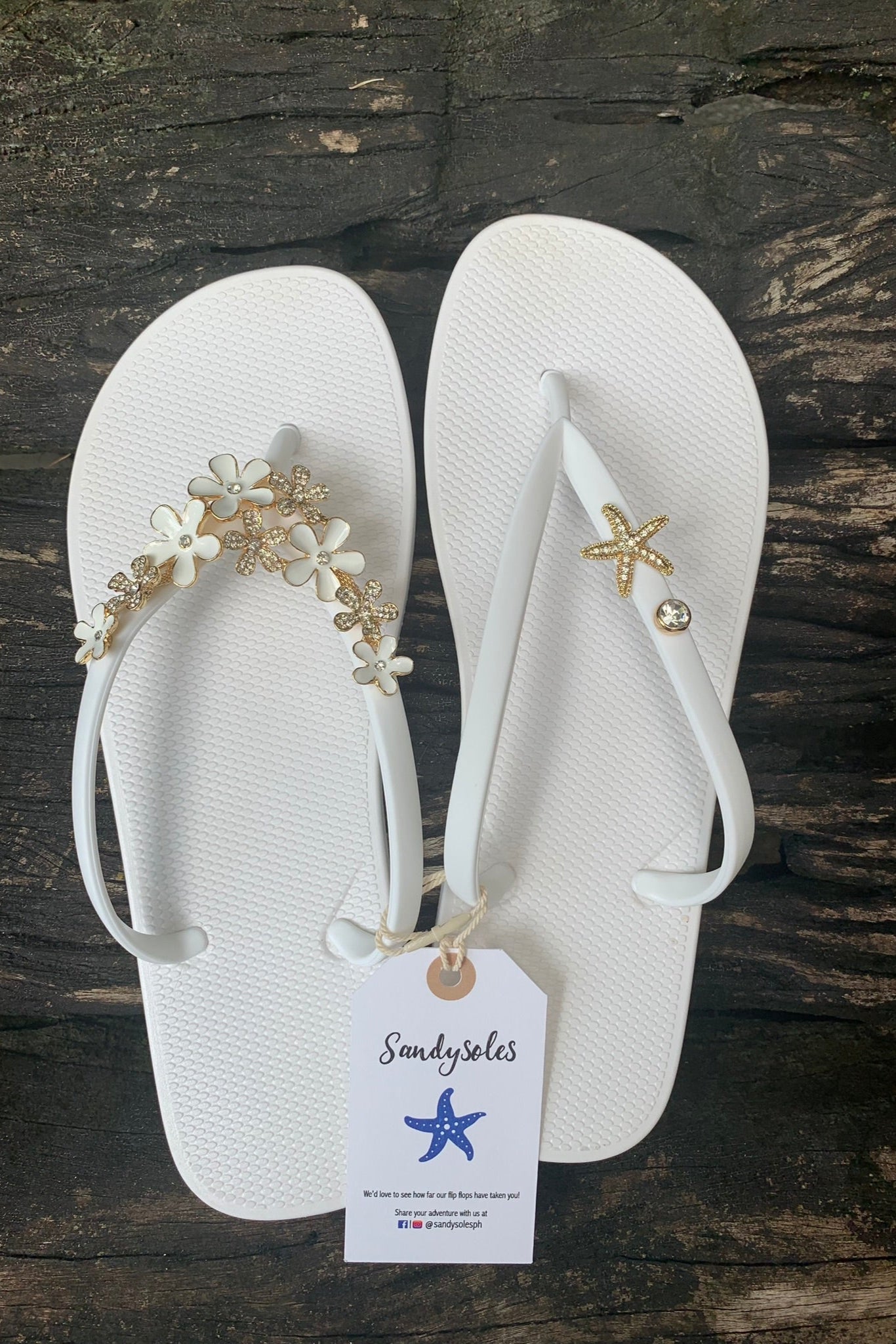 White flip-flops with decorative charms on straps, featuring a SandySoles tag.