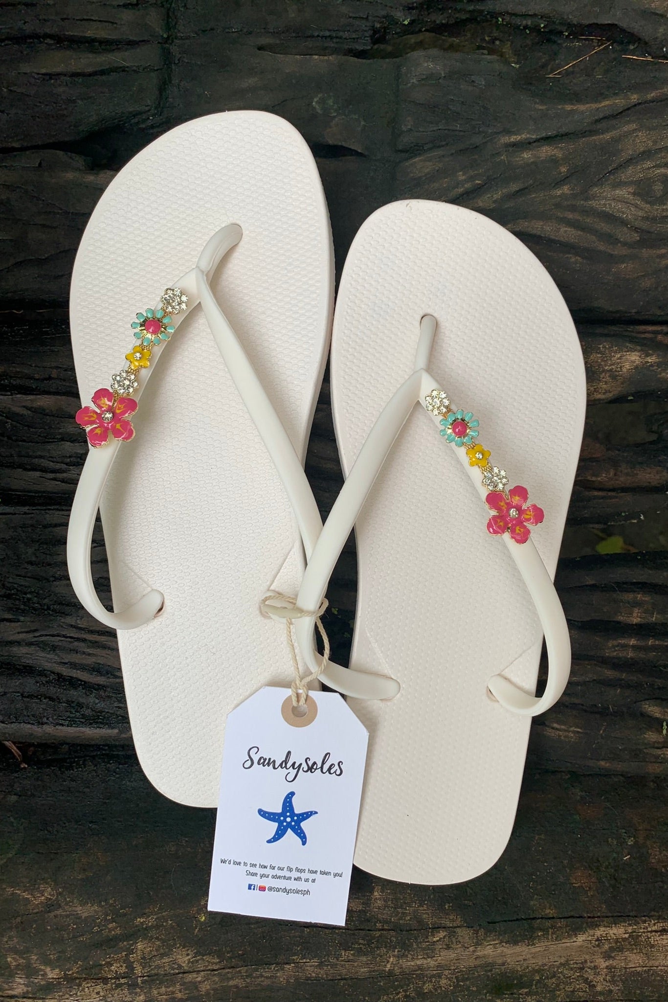 White flip-flops with pink flowerettes charm, featuring a 'Sandysoles' tag.