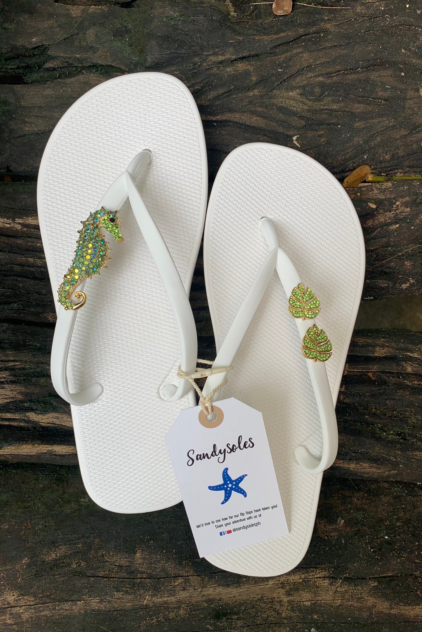White flip-flops with seahorse and monstera embellishments, featuring a SandySoles tag.