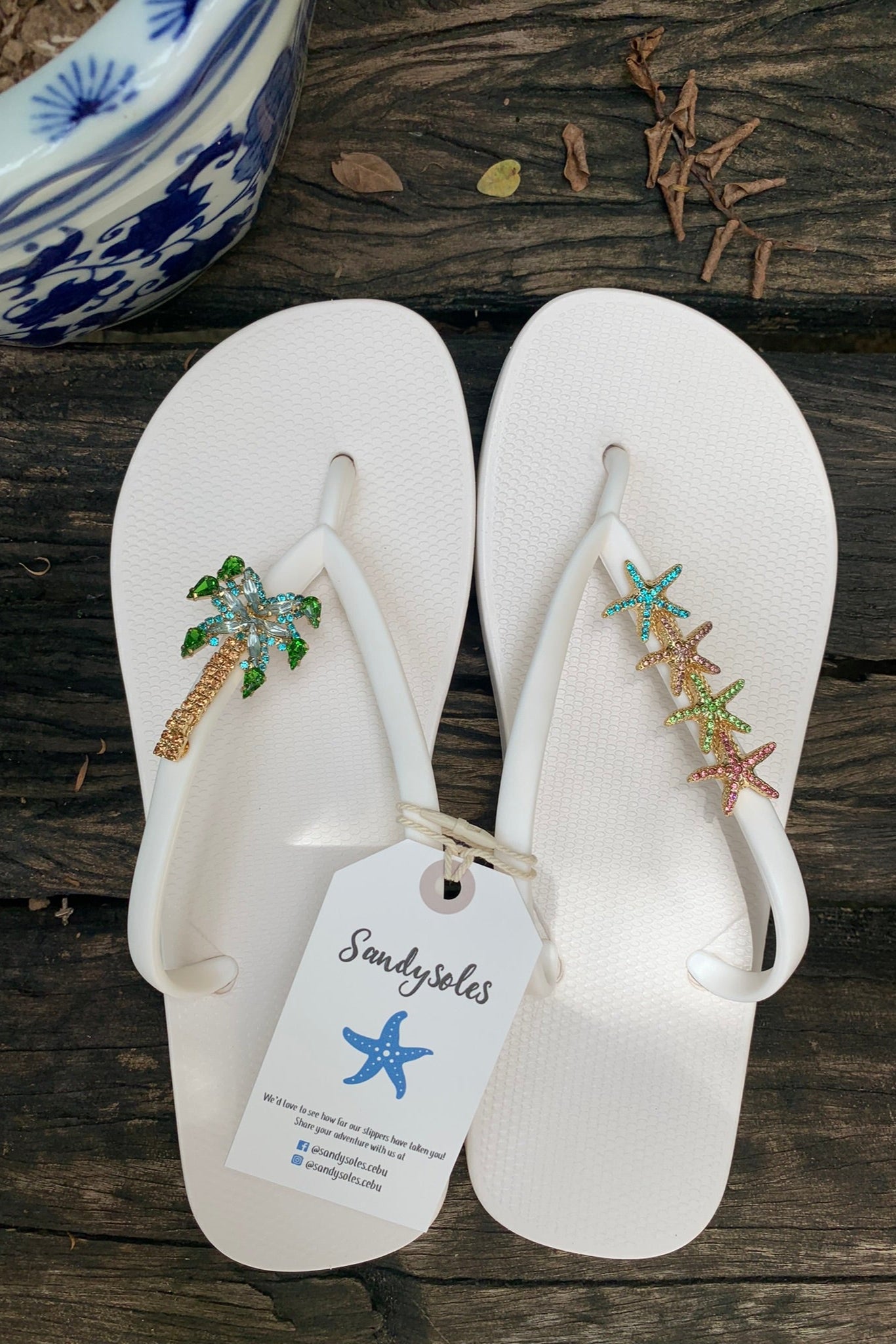 White flip-flops with decorative plam tree and starfish charms with a 'Sandysoles' tag.