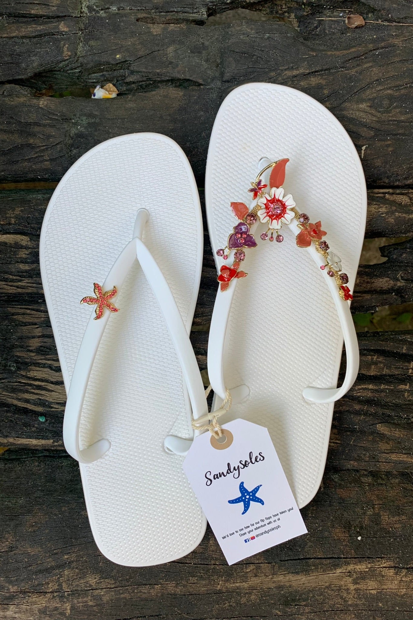 White flip-flops with decorative floral and starfish charms on straps, featuring a 'Sandysoles' tag.
