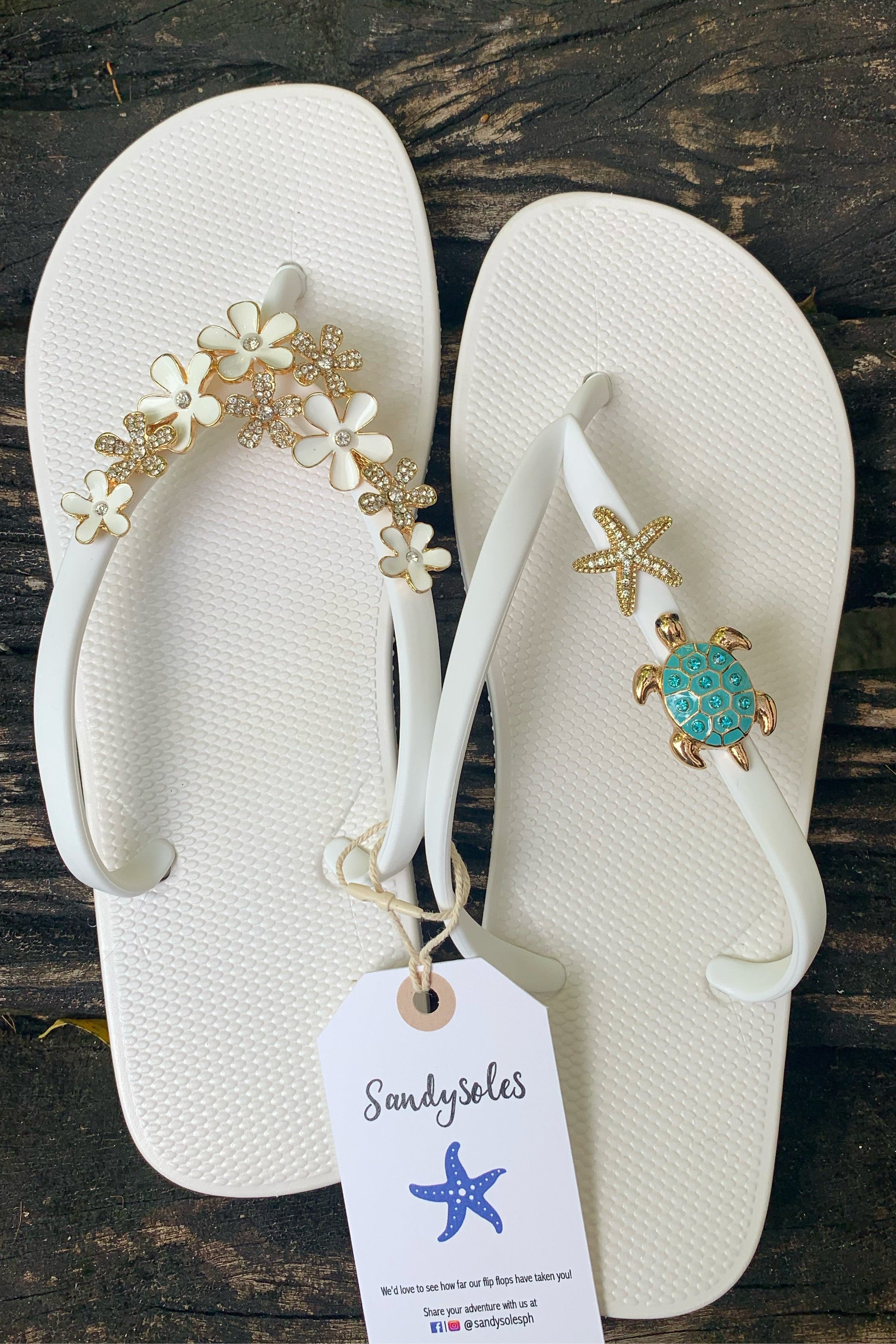 White flip-flops with decorative charms on straps, featuring a 'SandySoles' tag.