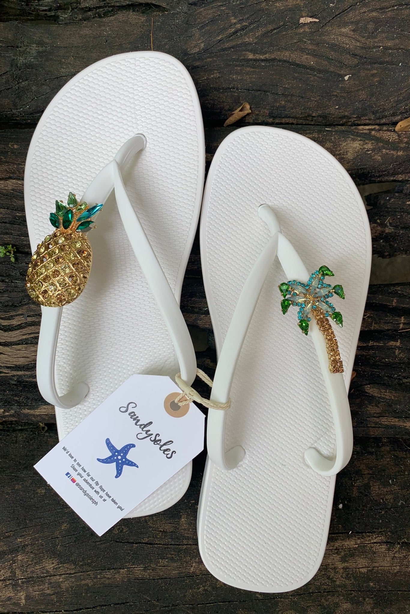 White flip-flops with pineapple and palm tree decorations featuring a 'Sandysales' tag.